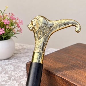 Könnte beinhalten: Ein Gehstock mit einem polierten, goldfarbenen Metallgriff in Form eines Löwenkopfes mit gebogenem Hals. Der Griff ist über eine glatte, zylindrische, goldfarbene Metallmanschette mit drei horizontalen Rillen mit einem dunkelbraunen Holzschaft verbunden.