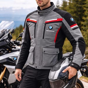Pode incluir: Casaco de motociclista cinza e preto com detalhes em vermelho e branco, com o logotipo da BMW. O casaco tem vários bolsos e uma gola alta. Uma motocicleta e um capacete também são visíveis.