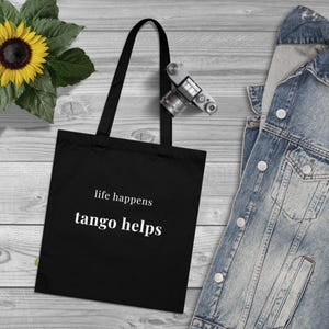 以下が含まれることがあります： 白い文字で「life happens tango helps」と書かれた黒いトートバッグ。バッグには長い黒いハンドルが付いており、明るい木製の表面に表示されています。ヒマワリとデニムジャケットも画像にあります。