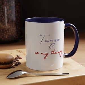 Puede incluir: Taza de cerámica blanca con interior y asa azul marino. La taza presenta el texto "Tango is my therapy" en escritura azul y roja. Una cuchara plateada y un macaron están sobre una superficie de madera.