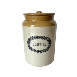 May include: A vintage ceramic jar with a lid, featuring a cream-colored base and a tan top. The jar is labeled "LENTILS" in black script within a floral wreath design, suggesting a kitchen storage container.