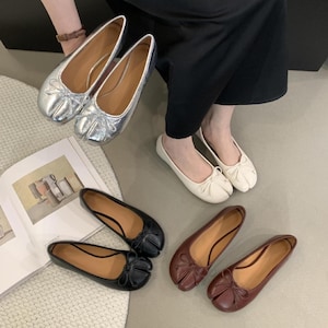 May include: A collection of ballet flats in various colors. The flats include silver, white, black, and burgundy. Each pair features a bow detail on the toe and a split-toe design. The shoes are displayed on a light-colored surface.