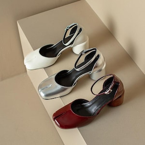 May include: Three pairs of heeled sandals in white, silver, and burgundy. Each sandal features a square toe, ankle strap, and a block heel. The shoes are displayed on a neutral-toned surface.