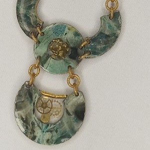 May include: A gold chain necklace with green and blue marbled pendant charms. The charms are shaped like half circles and feature a small gear mechanism.