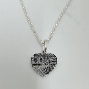 May include: A silver necklace with a heart-shaped pendant. The pendant features the word "LOVE" in a block letter style, with the letters encrusted with small, clear stones. The heart has a textured, radial design.