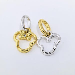 May include: Two charm pendants, one gold-toned with a flower and a small gem, the other silver-toned with the word "Better". Both pendants are shaped like a mouse head with a loop for attaching to a bracelet or necklace.