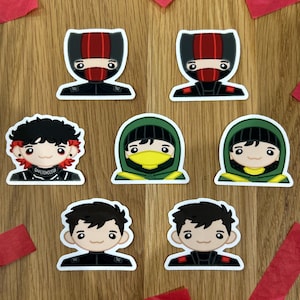 Könnte beinhalten: Eine Sammlung farbenfroher Aufkleber mit Cartoon-Charakteren in verschiedenen Outfits, darunter Ninja-ähnliche Kostüme und Kapuzenfiguren. Rotes Konfetti ist auf einer Holzoberfläche um die Aufkleber verstreut.