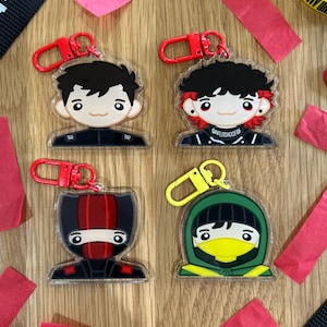 Könnte beinhalten: Vier Schlüsselanhänger aus Acryl mit Cartoon-Charakter-Designs. Jeder Schlüsselanhänger zeigt einen anderen Charakter mit schwarzen Haaren und Outfits, befestigt an einem roten oder gelben Verschluss. Rote Papierstreifen und schwarzes Band mit weißem Text sind verstreut.