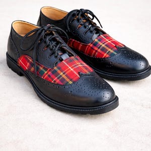 Op de afbeelding: Een paar zwarte leren Oxford-schoenen met rode, gele en groene geruite stoffen accenten. De schoenen hebben een klassiek wingtip-ontwerp met decoratieve perforaties en zwarte veters. Zwarte zolen.