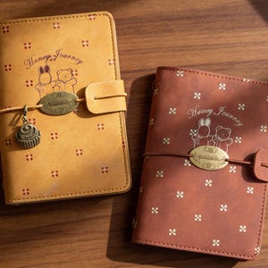 Puede incluir: Se muestran dos diarios de cuero, uno amarillo mostaza y otro marrón rojizo. Ambos presentan un patrón floral repetido y las palabras "Honey Journey" con una ilustración de un conejito y un oso. Cada diario tiene un cierre y un dije.