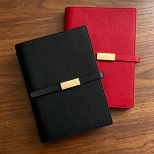 May include: Two leather-bound journals, one black and one red, are displayed on a wooden surface. Each journal features a strap with a gold-colored metal accent. The journals are closed and appear to be new.