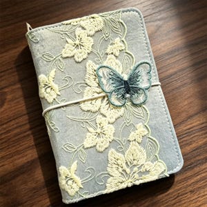 May include: A light blue fabric notebook cover featuring cream-colored floral embroidery across the front. A small, dark blue embroidered butterfly applique is attached to the center, held in place by a thin, light-colored elastic band.