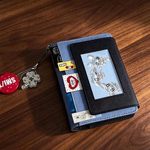 May include: A blue and black wallet featuring a clear ID window displaying three cartoon characters. The wallet is decorated with small rectangular stickers and includes a silver chain attachment with a red circular charm and a clover charm.
