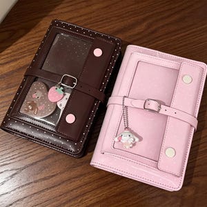 May include: Two rectangular binders with polka dot patterns, one dark brown and one light pink. Each features a clear front window, a buckled strap closure, two snap buttons, and a hanging charm: a pink strawberry on the brown and a white character on the pink.