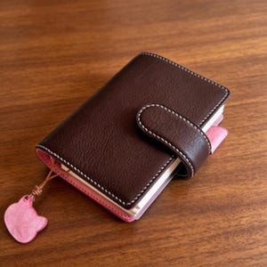 May include: A small, dark brown leather journal with a pink interior and a matching pink cat-shaped charm. The journal has a flap closure with white stitching and is closed. The journal is on a wooden surface.
