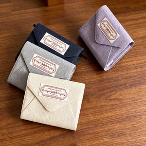 May include: Four small, square fabric wallets in shades of beige, gray, navy, and lavender. Each wallet has a flap closure and a rectangular label with text. The wallets are arranged on a wooden surface.
