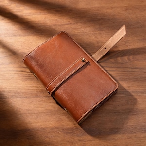 May include: A brown leather wallet with a strap closure. The wallet is rectangular with rounded corners and features visible stitching. The strap is secured with a gold-colored stud. The wallet is on a wooden surface.