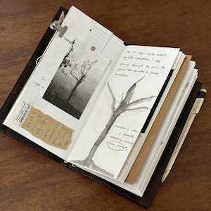 May include: Open leather journal with handwritten notes, sketches, and attached ephemera. The pages feature a black and white tree drawing, a vintage photo, and a sticker with Japanese text. A pen is tucked into the spine.