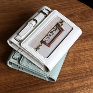 Puede incluir: Dos carteras de cuero, una blanca y otra verde claro, apiladas sobre una superficie de madera. La cartera blanca tiene una correa con hebilla. La cartera blanca tiene una ventana transparente con una imagen de un marco de madera y vegetación.