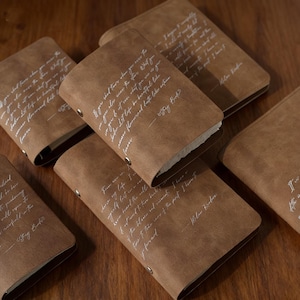 May include: A collection of brown leather-bound journals with white handwritten quotes. The journals are rectangular, with rounded corners and metal rivets. The pages are visible, and the overall aesthetic is rustic and vintage.