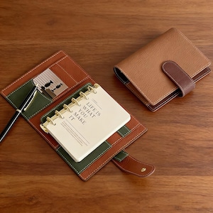 Könnte beinhalten: Ein braunes Leder-Notizbuch mit passendem Stift und einem kleinen Notizblock mit der Aufschrift "LIFE IS WHAT YOU MAKE IT". Das Notizbuch ist geöffnet und zeigt ein grünes und braunes Innere mit einer kleinen Karte. Ein geschlossenes braunes Leder-Notizbuch ist ebenfalls sichtbar.