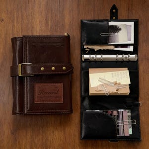 May include: A brown leather journal with a buckle closure and the brand name "Recaril" embossed on the front. The journal is next to a black leather organizer with a binder, paper clips, and small cards.