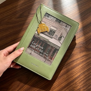 Op de afbeelding: Een saliegroen notitieboek met een rechthoekig venster op de kaft met een zwart-wit straatbeeld en de tekst Let green BREATHE slowly easing every daily tide. Een gouden auto-bedel hangt aan een kralenketting op de kaft.