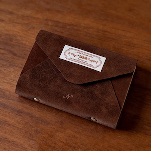 May include: A brown leather wallet in an envelope style with a flap closure. The wallet features a small white label with text and decorative elements. The letter "N" is embossed on the front. Two small metal rivets secure the flap.