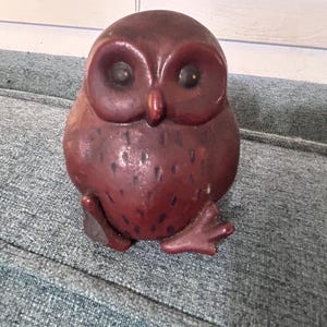 May include: A dark reddish-brown owl figurine with a smooth, rounded body, large circular eyes, and a small beak. The chest area features subtle, darker vertical markings, and the base shows two small, stylized feet extending outward.