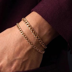 May include: Two gold-toned metal chain bracelets featuring interlocking flat links. The bracelets are worn stacked together, displaying a polished, reflective surface and a classic curb chain design that wraps around the wrist area.