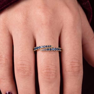 Könnte beinhalten: Ein goldener Bypass-Ring mit zwei überlappenden Bändern, die mit kleinen, dunkelblauen Steinen besetzt sind. Der Ring hat eine polierte Goldoberfläche und ein zartes, offenes Design, das sich leicht um den Finger legt.