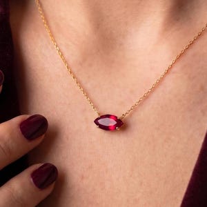 Peut inclure: Un collier à fine chaîne dorée orné d'un pendentif en pierre précieuse rouge foncé taillée en marquise. Le bijou repose sur le cou, mettant en valeur la pierre cramoisie éclatante fixée dans une monture métallique simple et fine.