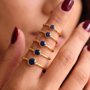 May include: Five gold rings, each featuring a single round, bezel-set blue sapphire gemstone. The rings are worn stacked along the length of a finger, showcasing a polished gold band and a deep blue stone in each setting, creating a uniform look.