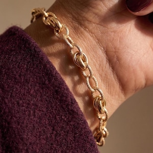 Puede incluir: Una pulsera de cadena dorada que presenta una serie de eslabones ovalados entrelazados. Cada eslabón individual muestra un acabado de superficie texturizado distintivo, creando un patrón sutil en el metal. La pulsera se lleva en la muñeca.