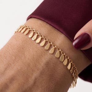 May include: A gold-toned chain bracelet featuring a series of small, uniform, teardrop-shaped charms dangling from each link. The bracelet is worn around the wrist, showcasing a delicate design with a polished metallic finish and consistent spacing.
