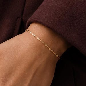 Może przedstawiać: Delikatna bransoletka z złotego łańcuszka z małymi, płaskimi, okrągłymi ogniwami, które odbijają światło. Bransoletka jest noszona na nadgarstku, spoczywając na rękawie ciemnobordowego płaszcza, prezentując prosty i wyrafinowany metalowy design.