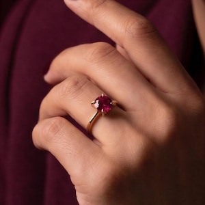 Op de afbeelding: Een gouden ring met een ronde, gefacetteerde, dieprode edelsteen in een zetting met vier pootjes. De ringband is dun en gepolijst, met een klassiek ontwerp dat de levendige kleur en helderheid van de centrale steen op de vinger benadrukt.