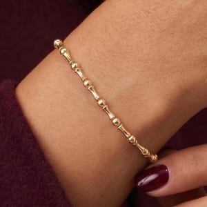 Può includere: Un delicato bracciale a catena in oro caratterizzato da un motivo ripetitivo di piccole perle sferiche lucide collegate da sottili barre metalliche dritte. Il bracciale ha una finitura metallica brillante e riflettente sul polso.