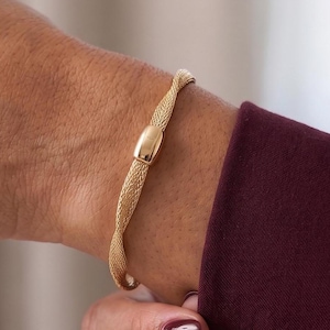 May include: A gold-toned bracelet featuring a twisted mesh band design with a smooth, polished rectangular center element. The bracelet has a flexible, woven texture that wraps around the wrist, showcasing a metallic finish and a refined, uniform structure.