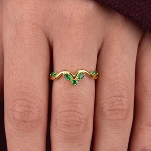 Könnte beinhalten: Ein goldener Ring in Chevron-Form mit fünf kleinen, runden, leuchtend grünen Edelsteinen, die entlang des V-förmigen Bandes gefasst sind. Der Ring hat eine polierte metallische Oberfläche und ein spitz zulaufendes, symmetrisches Design.