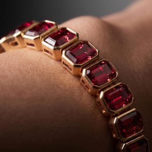May include: A gold bracelet featuring a series of rectangular, emerald-cut red gemstones set in individual gold bezels. The stones are arranged in a continuous line, each framed by a polished gold border, creating a linked chain design.