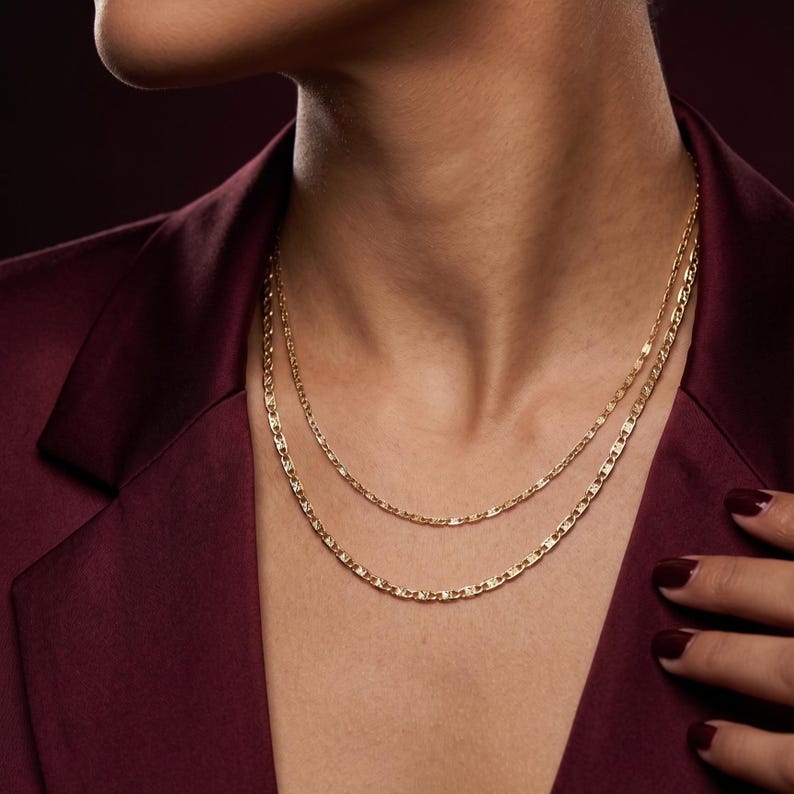 May include: Two gold-colored chain necklaces of different lengths are displayed. The necklaces have a delicate, flat link design. The person in the image is wearing a burgundy blazer. The background is a dark, solid color.