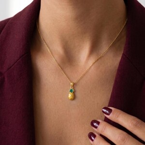 Può includere: Una collana dorata con un ciondolo a forma di ananas. Il ciondolo ad ananas ha un corpo dorato strutturato e una corona in smalto verde. La collana è indossata su un top bordeaux.