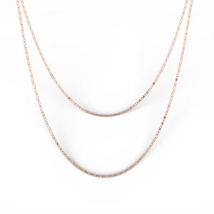 May include: Two-strand necklace in a rose gold tone. The delicate chains are composed of small, rectangular links. The necklace hangs against a white background, showcasing its simple elegance and design.