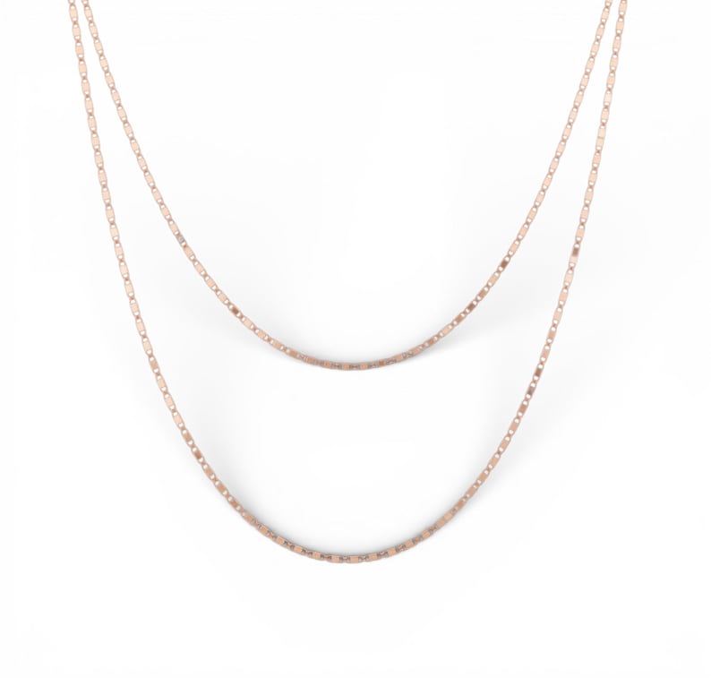 May include: Two-strand necklace in a rose gold tone. The delicate chains are composed of small, rectangular links. The necklace hangs against a white background, showcasing its simple elegance and design.