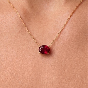 Op de afbeelding: Een delicate gouden ketting met een enkele ovaal geslepen dieprode robijn, gezet in een gouden zetting met vier pootjes. De hanger is gecentreerd aan een dunne, gepolijste gouden schakelketting die op de huid rust en de gefacetteerde steen toont.