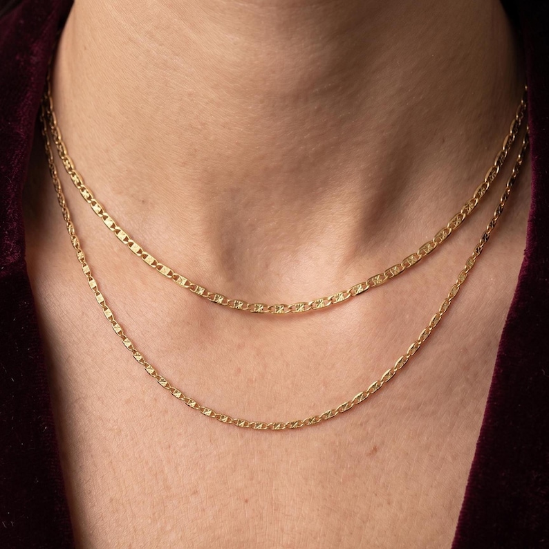 May include: Close-up of two gold necklaces with different chain styles. The top chain has a rectangular link design, while the bottom chain has an oval link design. Both necklaces are worn together, creating a layered look.