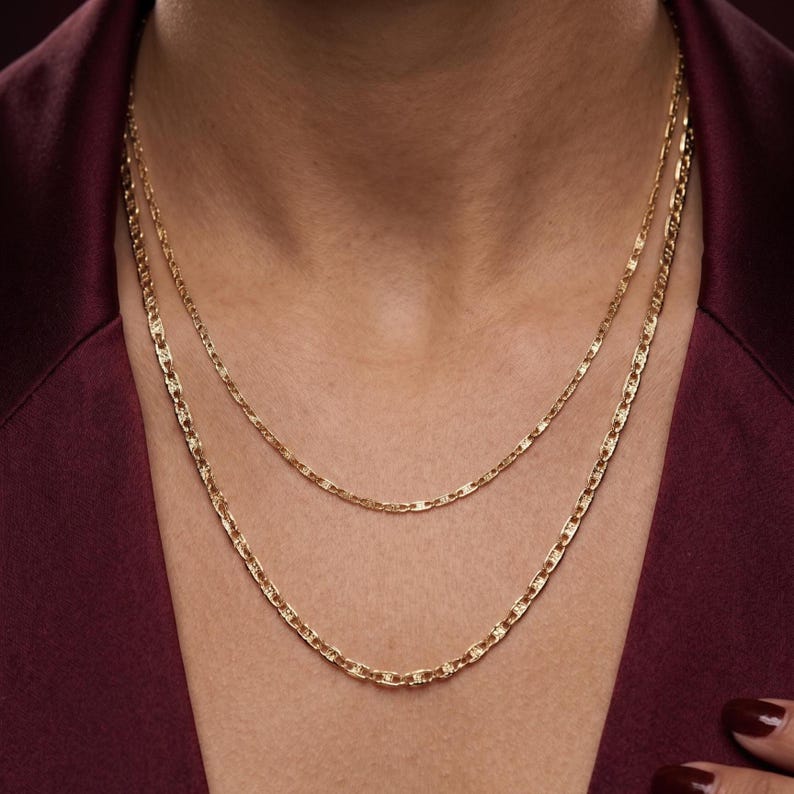 May include: Two gold-colored necklaces of different lengths. The necklaces have a delicate chain design. The background is a dark red color.