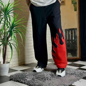 Puede incluir: Pantalones deportivos negros con un llamativo diseño de llamas rojas en la pierna derecha. Las llamas están delineadas en negro. Pantalones holgados, ideales para streetwear.