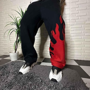 Könnte beinhalten: Schwarze Jogginghose mit rotem Flammendesign am rechten Bein. Die locker sitzende Hose hat einen Kordelzug in der Taille. Die Person trägt weiße und schwarze Turnschuhe.